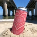 A pink heart-patterned beer can, "LoveStrong," standing in sand on a beach beneath a bridge.