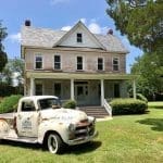 Vintage white pickup labeled "Hopped Up Farm Truck" parked before a two-story farmhouse with a front porch.