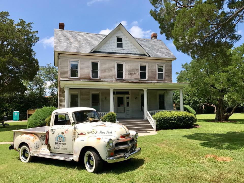 Vintage white pickup labeled "Hopped Up Farm Truck" parked before a two-story farmhouse with a front porch.