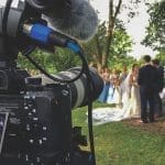 A video camera with a furry microphone records an outdoor wedding ceremony, with a bride in a white gown and bridesmaids in blue.