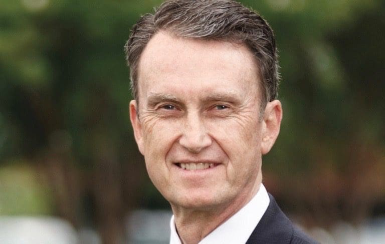 John T. Midgett smiles in a dark suit and white shirt before a blurred green tree background.