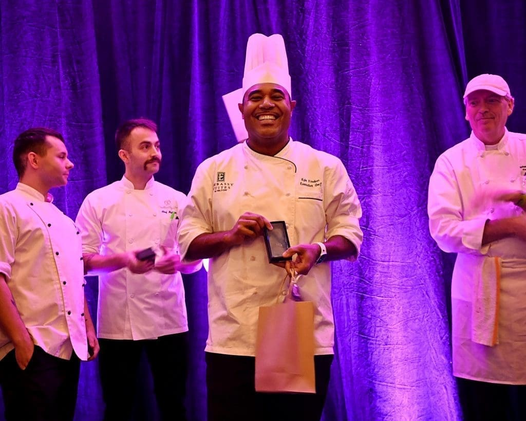 Four chefs in white coats stand on a stage before purple drapes, and the center chef smiles while holding an award plaque.
