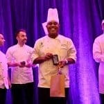 Four chefs in white coats stand on a stage before purple drapes, and the center chef smiles while holding an award plaque.