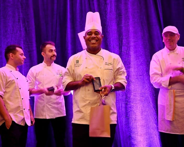 Four chefs in white coats stand on a stage before purple drapes, and the center chef smiles while holding an award plaque.