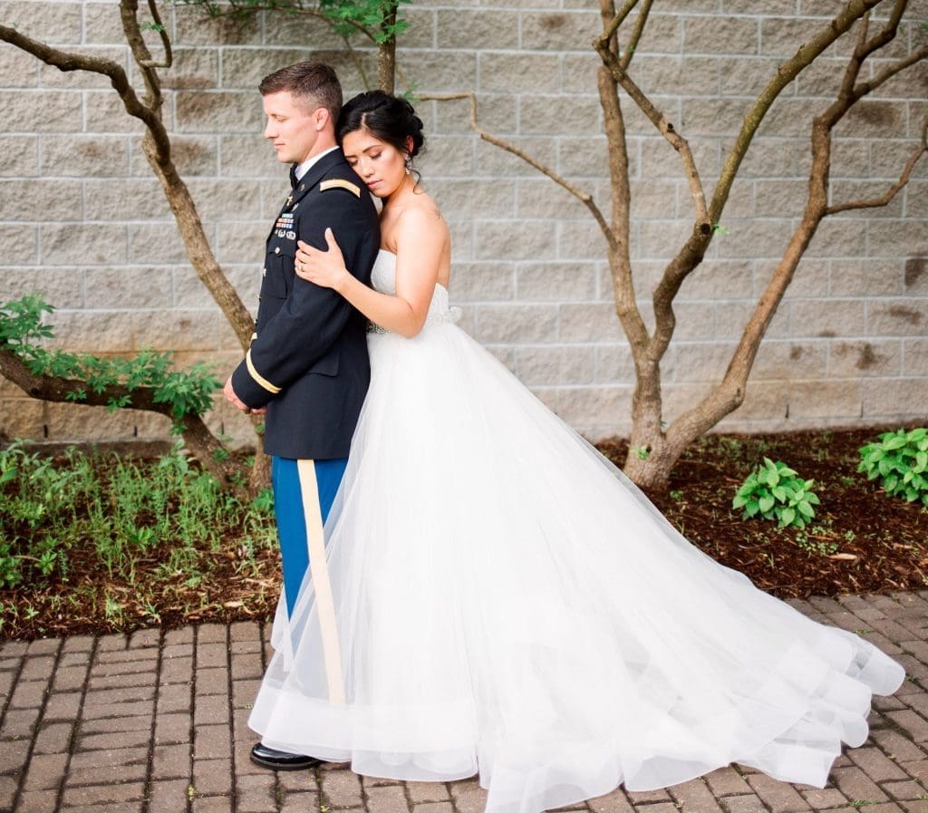A bride in a strapless tulle gown leans against a groom in a dark military dress uniform, standing back to back.