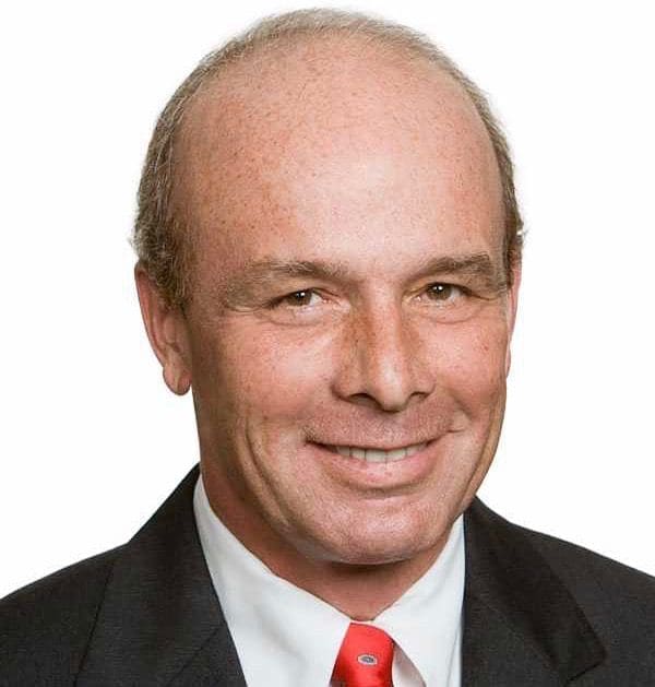 Smiling man with receding gray hair, wearing a dark suit, white shirt and red tie against a white background.