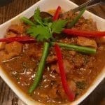 A white square bowl of chicken curry in rich brown sauce, garnished with cilantro, red pepper strips and green beans.