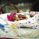 A tiny premature baby in a pink knit hat rests in a NICU incubator, with feeding tubes and monitor wires attached.