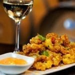 Crispy glazed bites garnished with cilantro, served with orange dipping sauce beside a glass of white wine.