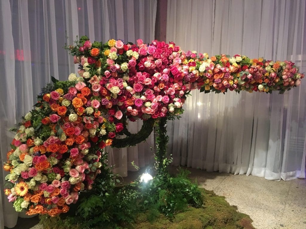 A large sculpture of a chameleon covered in pink, orange, and white roses, set on a moss-covered floor before white drapes.