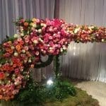 A large sculpture of a chameleon covered in pink, orange, and white roses, set on a moss-covered floor before white drapes.