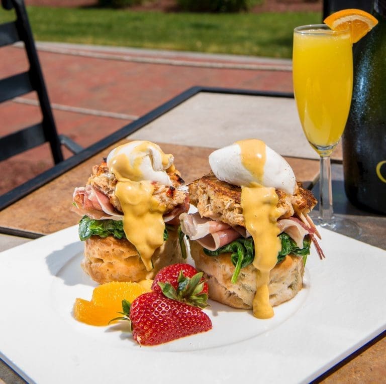 Two crab cake eggs Benedict biscuits with hollandaise, spinach and ham on a white plate with strawberries, beside a mimosa.