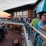 Patrons crowd a waterfront restaurant's upper deck at sunset, overlooking the harbor with a red umbrella and turquoise chairs.