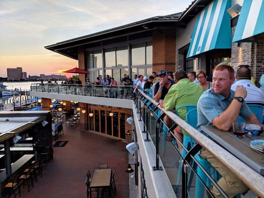Patrons crowd a waterfront restaurant's upper deck at sunset, overlooking the harbor with a red umbrella and turquoise chairs.