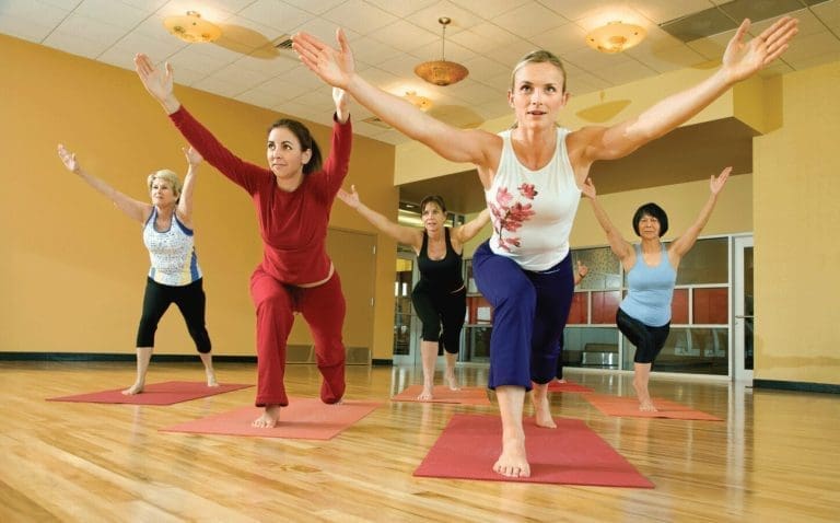Five women in a bright studio hold warrior poses on red mats, arms raised overhead.