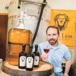 A bearded man in a blue polo holds a glass of mead beside bottles and a glass carboy on a barrel.