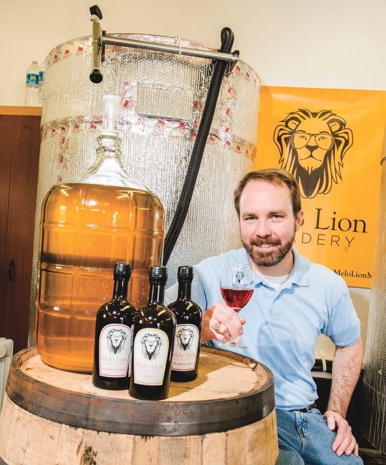 A bearded man in a blue polo holds a glass of mead beside bottles and a glass carboy on a barrel.