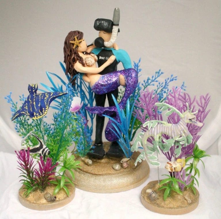 A sculpted clay cake topper of a diver in a wetsuit embracing a mermaid with a purple glitter tail, surrounded by sea creatures and coral.