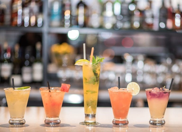 Five colorful cocktails in tall and short glasses sit in a row on a bar, garnished with lime, watermelon and mint.