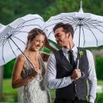 A smiling bride in a beaded gown and a groom in a gray vest share two white umbrellas outdoors.