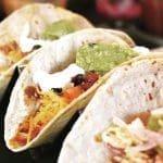 Three soft flour tacos topped with guacamole, sour cream, shredded cheese, and diced tomato in a metal holder.
