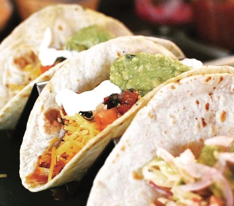 Three soft flour tacos topped with guacamole, sour cream, shredded cheese, and diced tomato in a metal holder.