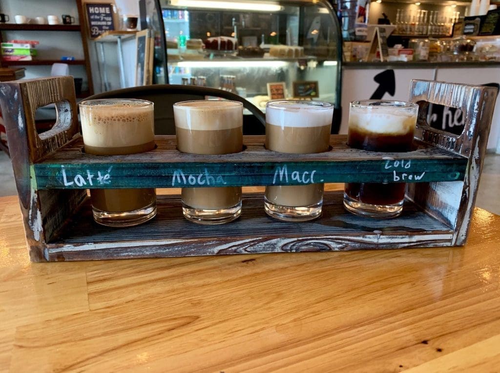A wooden flight tray holds four small glasses of coffee labeled Latte, Mocha, Macchiato, and Cold Brew.