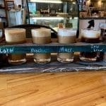 A wooden flight tray holds four small glasses of coffee labeled Latte, Mocha, Macchiato, and Cold Brew.