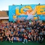 Large crowd of people posing in front of a colorful LOVE mural, holding a black NERC banner and trophy.