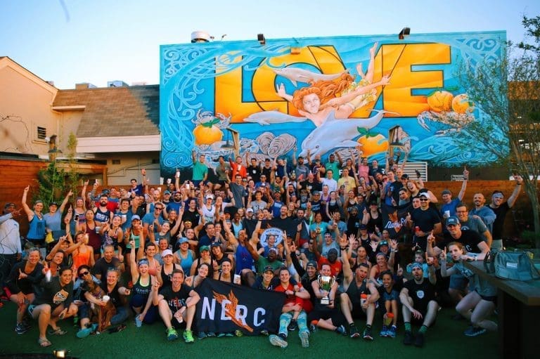 Large crowd of people posing in front of a colorful LOVE mural, holding a black NERC banner and trophy.