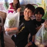 Smiling children hold plastic grocery bags filled with carrots, greens and other produce in front of a painted mural.