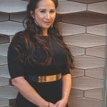 Jeannette Rincon stands before geometric wood-tone wall panels, wearing a black dress with a gold waist belt.