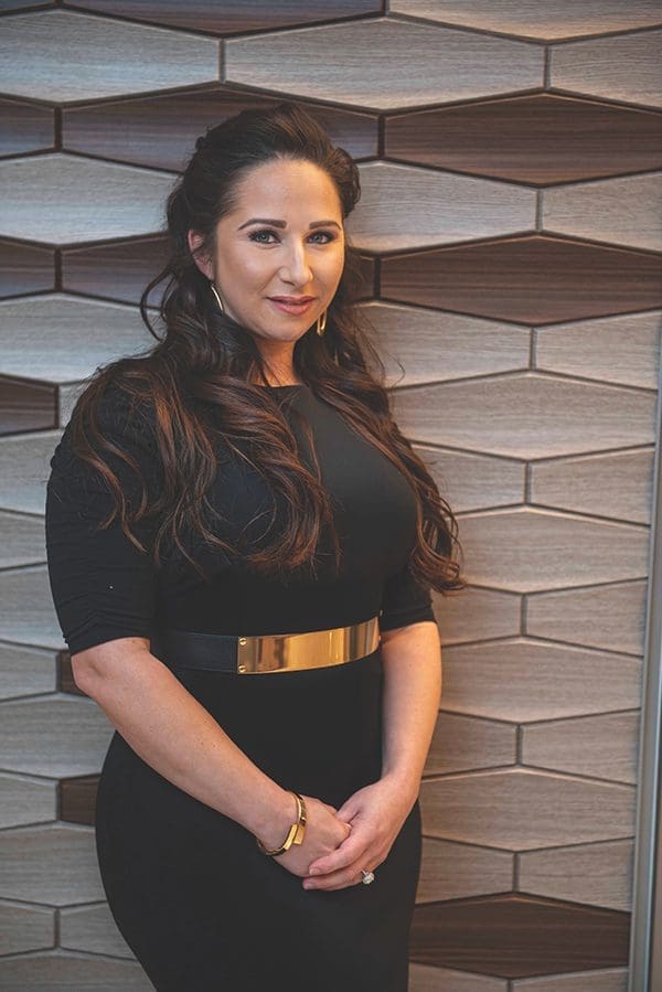 Jeannette Rincon stands before geometric wood-tone wall panels, wearing a black dress with a gold waist belt.