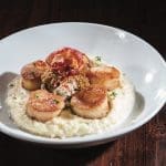 Seared scallops with crispy prosciutto, mustard seeds, and herbs over creamy white grits on a white plate.