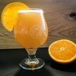 A tulip glass of cloudy orange-gold beer with foam, garnished with an orange slice, beside a halved orange.