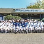 A large group of chefs in white uniforms and toques poses outside the Culinary Institute of Virginia building and a branded truck.