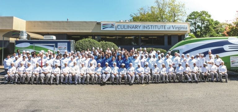 A large group of chefs in white uniforms and toques poses outside the Culinary Institute of Virginia building and a branded truck.
