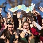 A crowd of smiling young people raise hands toward a paper caterpillar cutout against a blue sky and bare trees.