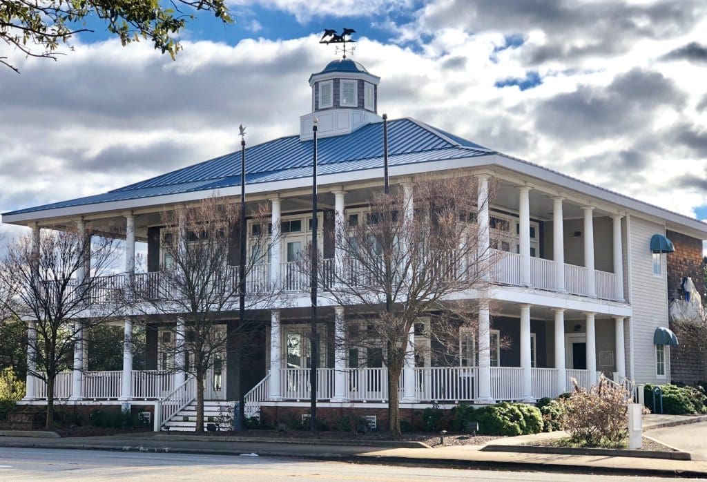 A two-story white colonial-style building with a wraparound porch, blue metal roof, and cupola with weathervane.