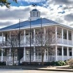 A two-story white colonial-style building with a wraparound porch, blue metal roof, and cupola with weathervane.
