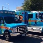 Two bright blue pediatric ambulances with orange stripes and colorful sea creature artwork parked on a sunny street.