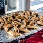 Silver tray of golden fried spring rolls garnished with chopped chives, set on a red cloth at an outdoor event.