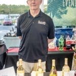 A smiling man in a dark polo stands behind a table of white wine bottles, with a "Wine Provided By" sign.