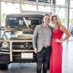 A smiling man in a gray blazer and a woman in a red dress pose beside a matte black Mercedes-Benz G-Class SUV in a dealership showroom.