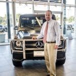 A man in a light shirt, red tie and khakis stands beside a matte black Mercedes-Benz G-Class SUV in a showroom.