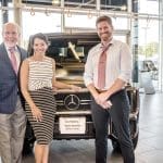 Three smiling people pose beside a dark Mercedes-Benz G-Class SUV in a dealership showroom.