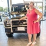 A smiling woman in a coral dress poses beside a matte black Mercedes-Benz G-Class SUV in a dealership showroom.