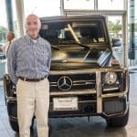 A smiling man in a checked button-down and khakis stands beside a black Mercedes-Benz G-Class SUV in a showroom.
