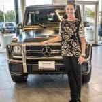 Smiling woman in a floral top poses beside a matte black Mercedes-Benz G-Class SUV in a showroom.