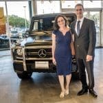 Two people smile beside a black Mercedes-Benz G-Class SUV inside a glass-walled dealership showroom.